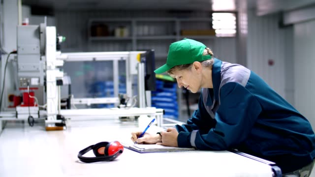 elderly-woman,-employee,-worker-at-an-enterprise,-factory,-fills-up-a-service-journal,-record-book,-working-industrial-background