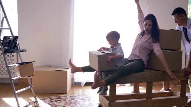 happy-relocation,-young-man-carries-wife-with-little-son-on-table-in-new-apartment-with-of-boxes-with-things
