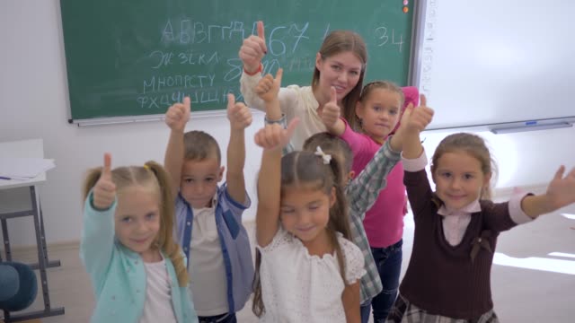 Retrato-de-la-escuela-a-los-niños-con-el-maestro-mostrando-los-pulgares-para-arriba-y-luego-aplaudir-en-aula-sobre-fondo-de-junta-en-la-escuela