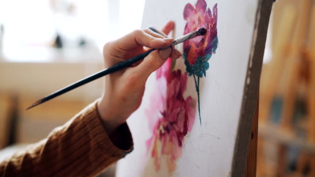 Close-up-shot-of-female-hand-holding-paintbrush-and-painting-bright-flowers-on-canvas-in-light-studio.-Pictorial-art,-artistic-work-and-people-concept.