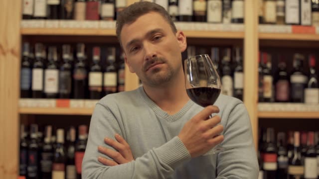 Portrait-of-handsome-man-drinking-red-wine.-Man-shakes-his-drink,-raises-wine-glass-and-takes-a-sip-of-wine-with-pleasure.-The-guy-with-the-wine-glass-on-the-background-of-the-shelves-with-wine-bottles.
