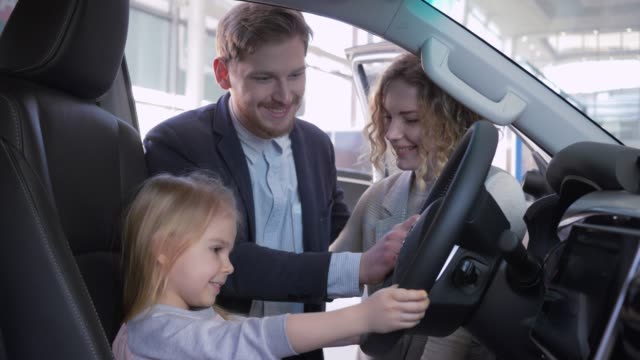 buying-family-car,-little-child-girl-behind-wheel-of-new-auto-together-with-parents-while-purchase-automobile-at-showroom-close-up
