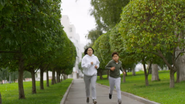 Sportliche-asiatische-Mutter-und-Sohn-Joggen-entlang-Green-Alley-mit-Bäumen