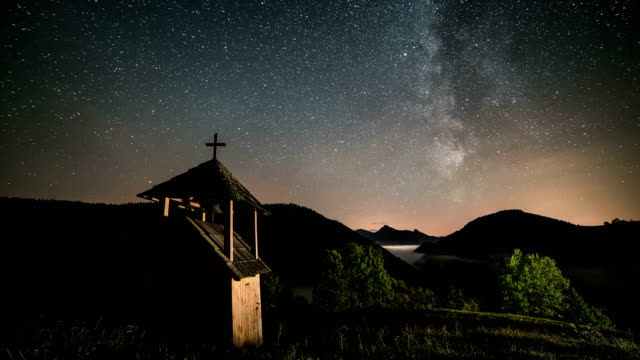 Stars-with-milky-way-galaxy-moving-over-wooden-belfry-in-mountains-and-foggy-valley-rural-country.-Dolly-shot-time-lapse-starry-night-sky