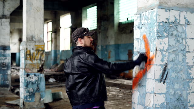 Bearded-graffiti-artist-is-painting-on-pillar-in-old-abandoned-building-using-aerosol-paint.-Modern-youth-subculture,-creative-people-and-street-art-concept.