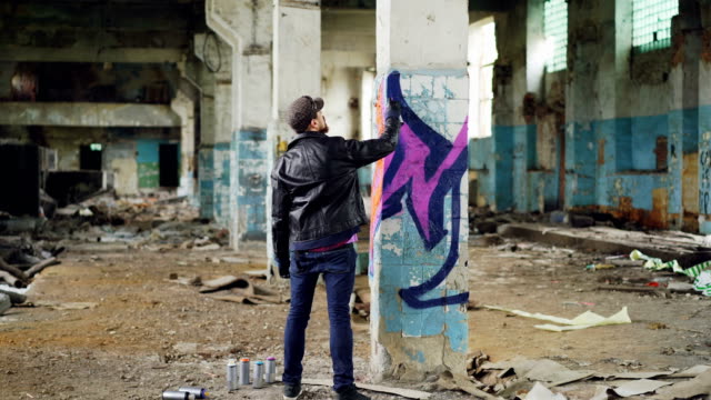 Graffiti-Maler-in-Freizeitkleidung-zieht-mit-Sprühfarbe-auf-Spalte-in-geräumigen-verlassenen-Gebäude.-Abstrakte-Bilder,-moderne,-kreative-Menschen-und-hipster-Konzept.