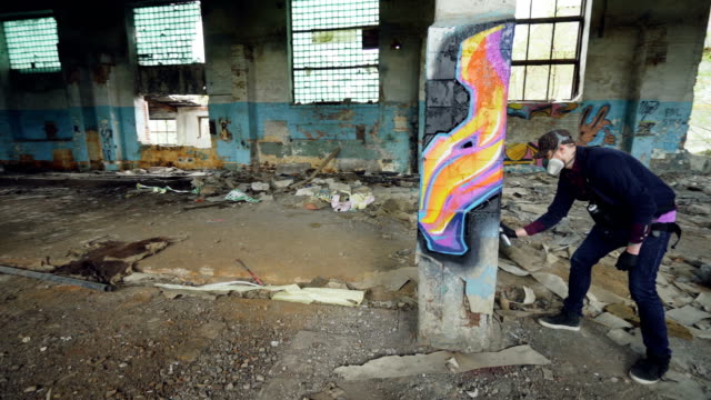 Pan-shot-of-masked-graffiti-artist-drawing-abstract-images-on-pillar-in-large-empty-building-using-paint-spray.-Painter-is-wearing-casual-clothing-and-protective-gloves.