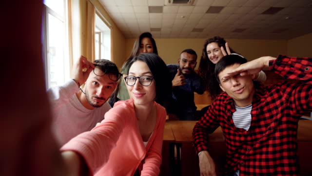 Point-of-view-shot-of-attractive-young-men-and-women-taking-selfie-in-lecture-hall,-posing-with-textbook-and-showing-hand-gestures-and-expressive-faces.