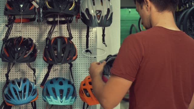 A-young-Caucasian-man-stands-near-a-stand-in-a-bicycle-store-in-the-head-protection-department.-Choosing-a-bicycle-helmet-in-a-small-store