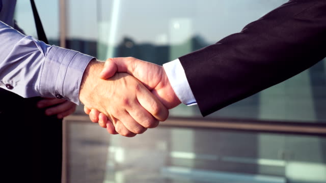 Close-up-of-successful-businessmen-greeting-each-other-near-office-building.-Two-young-colleagues-meeting-and-shaking-hands-in-urban-environment.-Handshake-of-business-partners-outdoor.-Side-view