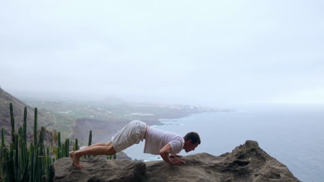 The-man-standing-on-the-edge-of-a-cliff-in-the-pose-of-the-dog-with-views-of-the-ocean,-breathe-in-the-sea-air-during-a-yoga-journey-through-the-Islands