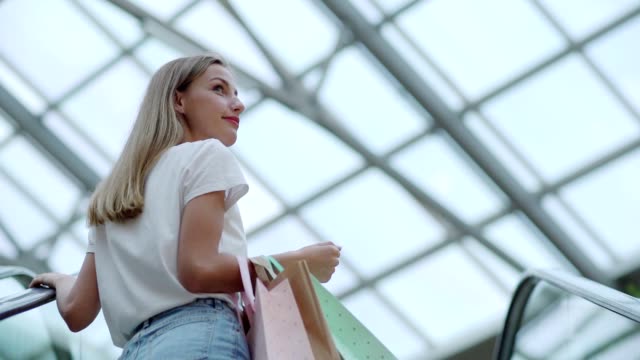 Atractiva-joven-con-bolsas-de-observar-el-centro-comercial-y-sonriendo-mientras-se-mueve-hacia-arriba-en-escalera-mecánica-en-movimiento-lento