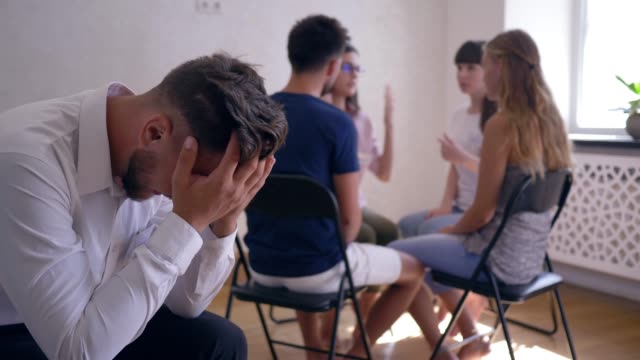 trauriger-Mann-weint-und-Abdeckungen-Gesicht-mit-den-Händen-auf-Gruppentherapie-Sitzung-auf-Hintergrund-von-Menschen-sitzen-auf-Stühlen-im-Kreis