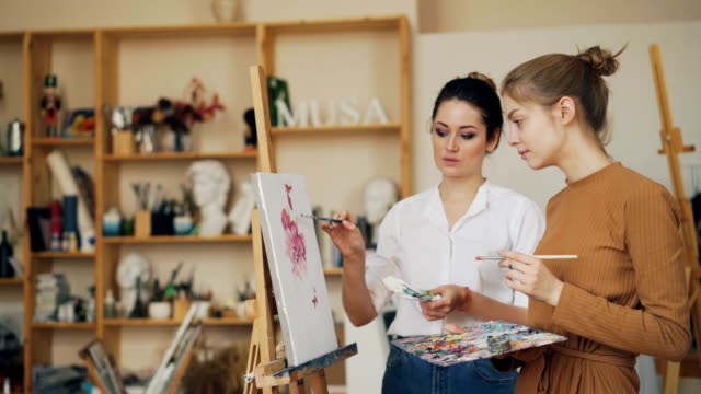 Experienced-painting-teacher-is-talking-to-her-female-student-standing-near-picture-on-easel-and-discussing-artwork.-Shelves-with-tools-are-visible.
