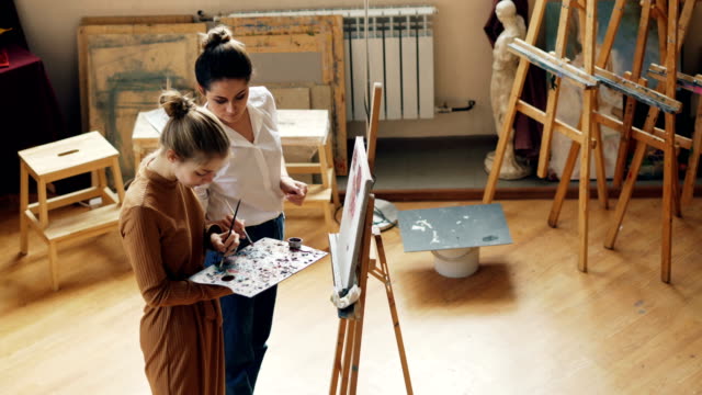 Alto-ángulo-de-visión-de-dos-mujeres-jóvenes-arte-maestra-y-alumno-mezclando-colores-en-la-paleta,-hablando,-sonriendo-y-pintura-sobre-lienzo-en-estudio-de-Artes-modert.