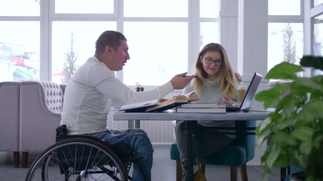 individuelles-Studium-für-Menschen-mit-Behinderungen,-Lehrerin-Frau-in-Brillen-führt-Vortrag-für-ungültige-männlich-am-Rollstuhl-mit-einem-Laptop-Computer-und-Bücher