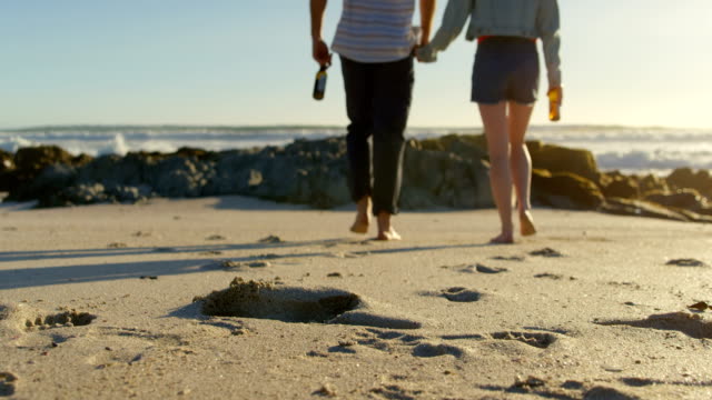 Couple-holding-hands-with-beer-bottle-walking-on-beach-4k