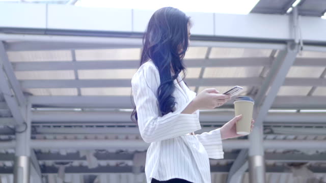 Junge-asiatische-Geschäftsfrau-mittels-Smartphone-laufen-und-trinkt-Kaffee-im-Stadt-Zentrum-Thailand.--Kommunikation,-Alltag-Technologiekonzept.-SLOW-MOTION