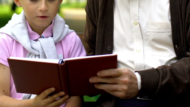 Abuelo-enseñando-a-nieto-leyendo,-descansando-sobre-un-banco-en-el-parque-de-verano,-familia