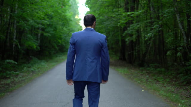 handsome-young-businessman-walking-along-the-road-in-forest,-view-back