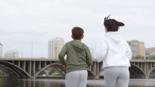 Rear-View-of-Mother-and-Son-Jogging-along-Riverside-in-City