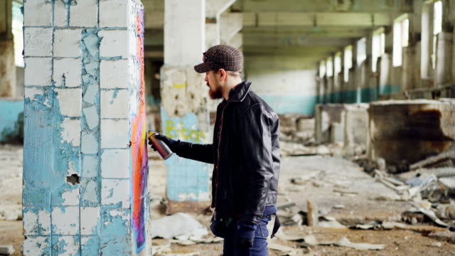 Side-view-of-graffiti-artist-bearded-guy-drawing-on-damaged-column-inside-emply-industrial-building-using-bright-aerosol-paint.-Creativity-and-people-concept.