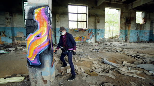 Young-man-professional-graffiti-painter-is-working-inside-abandoned-building,-he-is-painting-with-spray-aerosol-paint-on-high-column.-Modern-art-and-creativity-concept.