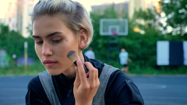 Close-footage-of-beautiful-female-basketball-player-applying-make-up-and-sitting-on-ground,-men-playing-in-background