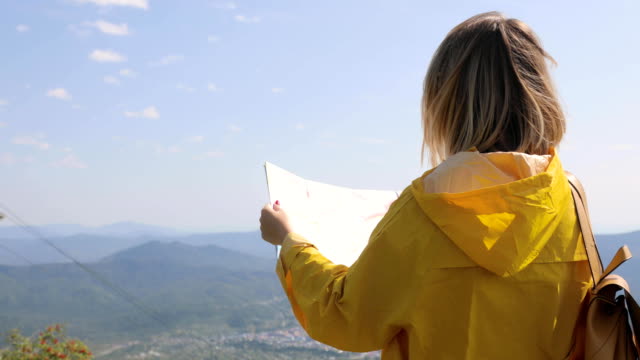 Back-view-of-Caucasian-female-hiker-in-yellow-raincoat-stands-in-the-mountains-with-a-map-in-hand