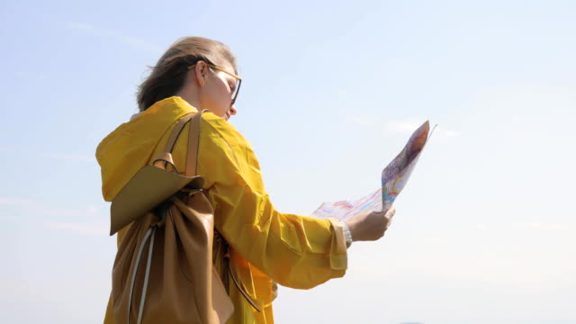 Back-view-of-Caucasian-female-hiker-in-yellow-raincoat-stands-in-the-mountains-with-a-map-in-hand
