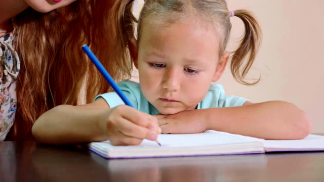 Little-cute-girl-sits-in-classroom-and-studies-with-teacher-in-exercise-book
