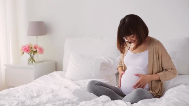 happy-pregnant-asian-woman-sitting-on-bed-at-home