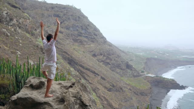 A-man-standing-on-the-edge-of-a-cliff-overlooking-the-ocean-raises-his-hands-up-and-inhales-the-sea-air-during-yoga