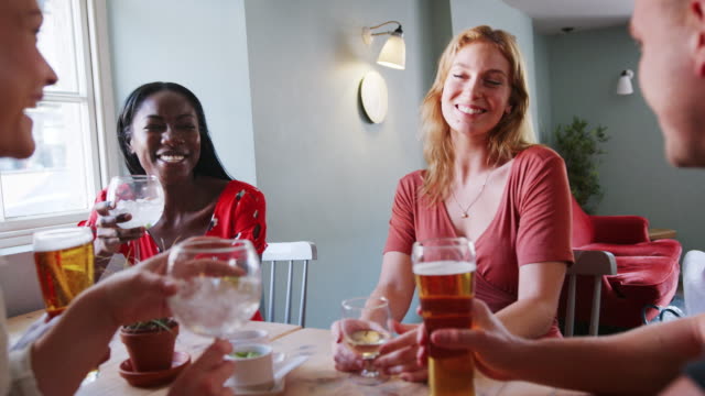 Cinco-jóvenes-amigos-adultos-sentados-con-bebidas-en-una-mesa-en-un-bar-hablando-y-riendo,-de-cerca,-bajo-ángulo