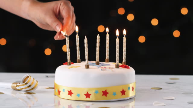 Close-up-of-a-hand-using-a-match-to-light-seven-candles-on-a-white,-decorated-birthday-cake,-a-party-blower-beside-it,-bokeh-lights-in-the-background