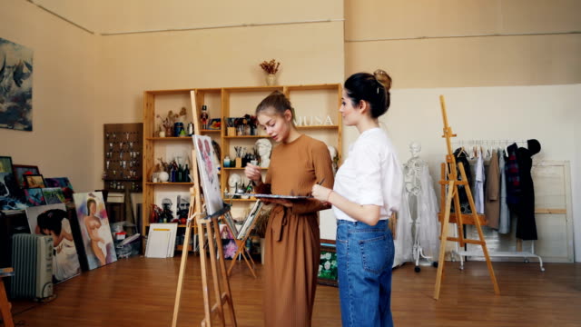 Cheerful-lady-is-painting-picture-together-with-her-female-teacher-professional-artist-using-brush-and-oil-paints.-Modern-workshop-with-easels-and-artworks-is-visible.