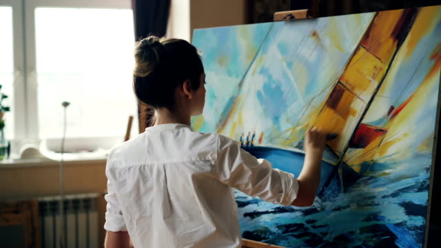 Serious-female-artist-is-working-at-picture-using-oil-paints-and-palette-knife-creating-beautiful-marine-landscape-on-canvas.-Painting-technique-and-painters-concept.