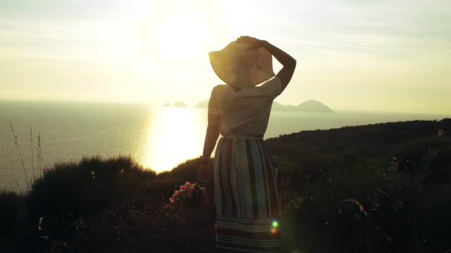 Schöne-junge-Frau-tragen-bunte-Kleid-Mode-mit-Rock-und-Hut-Stand-mit-Blumen-im-Korb-bei-Sonnenuntergang-auf-der-italienischen-Insel-Ponza-Berg-Küste.