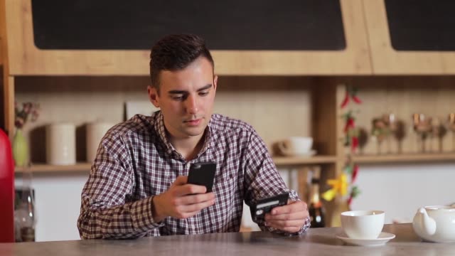 Handsome-man-making-purchase-online,-typing-credit-card-details-on-black-smartphone-in-the-modern-kitchen