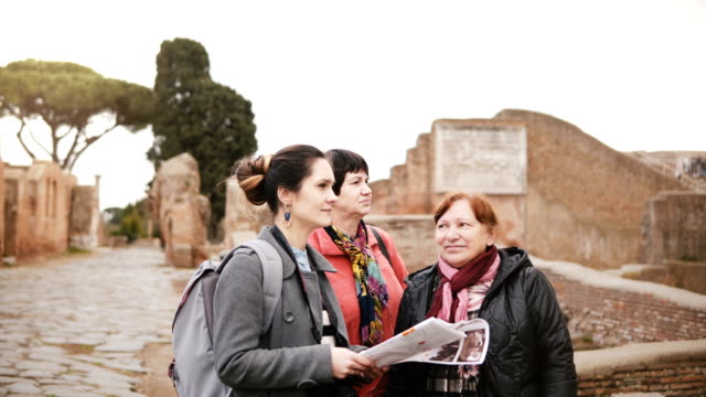 Dos-caucásicos-senior-femenino-viajeros-y-guía-mujer-joven-disfrutan-explorando-ruinas-históricas-de-Ostia,-Italia-de-vacaciones.