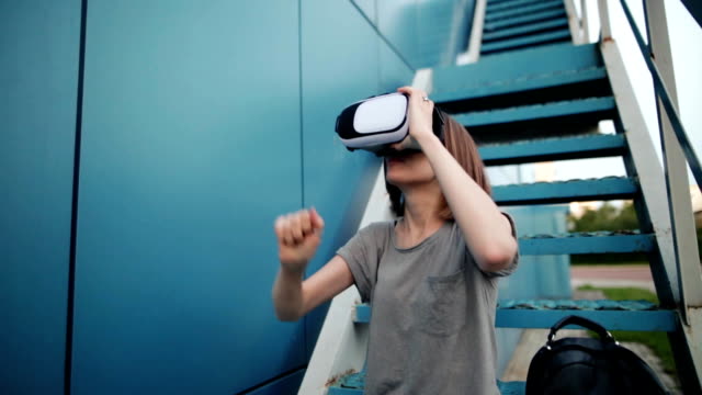 Zukunft-ist-jetzt.-Schöne-junge-Frau-auf-einer-Treppe-Rollenspiel-in-Vr-Brille.-Kaukasische-Mädchen-berühren-etwas-mit-modernen-virtual-Reality-Brille-auf-blauem-Grund.