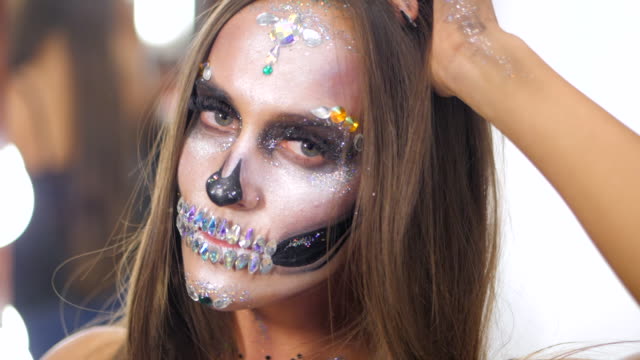 Scary-young-girl-with-creative-halloween-war-paint-in-the-dressing-room.-Portrait-of-glamorous-skull-with-rhinestones-and-sequins.-Professional-make-up-for-the-celebration.-Slow-motion