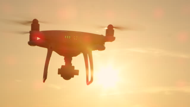 SLOW-MOTION-CLOSE-UP-SILHOUETTE:-Drone-flying-over-the-sun-on-golden-sunset-sky