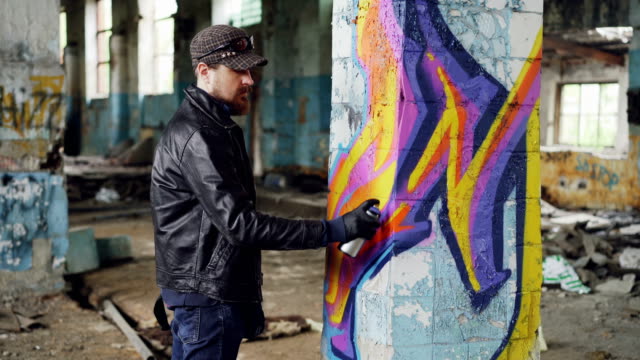 Professional-graffiti-painter-is-creating-abstract-image-on-large-pillar-inside-abandoned-building-with-spray-paint.-Young-man-is-wearing-leather-jacket,-cap-and-gloves.