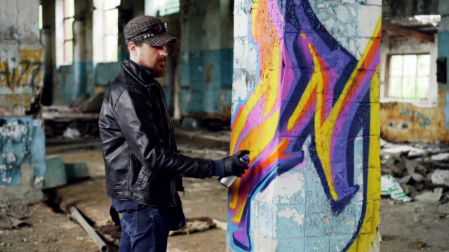 Professionelle-Graffiti-Maler-schafft-abstraktes-Bild-auf-große-Säule-in-verlassenen-Gebäude-mit-Sprühfarbe.-Junge-Mann-trägt-Lederjacke,-Mütze-und-Handschuhe.