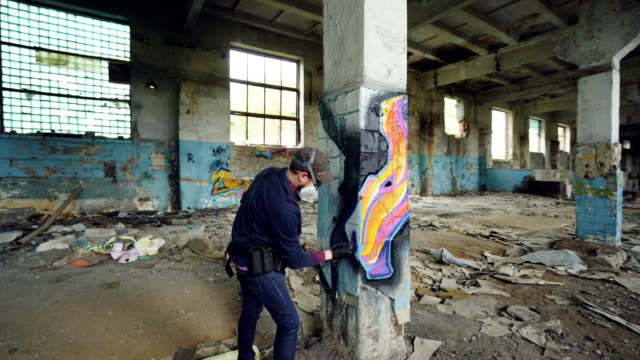 Pan-shot-of-adult-man-graffiti-artist-in-protective-face-mask-and-gloves-painting-on-high-pillar-inside-damaged-empty-industrial-building.-Creativity-and-people-concept.