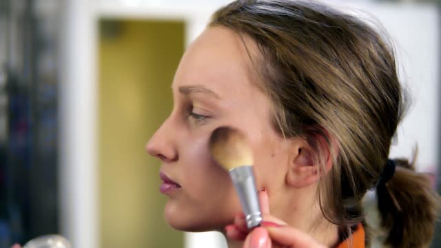 Side-view-of-a-young,-pretty-female-model.-Make-up-process.-Artist-putting-some-transparent,-shiny-rouge-on-woman's-cheek-using-a-brush