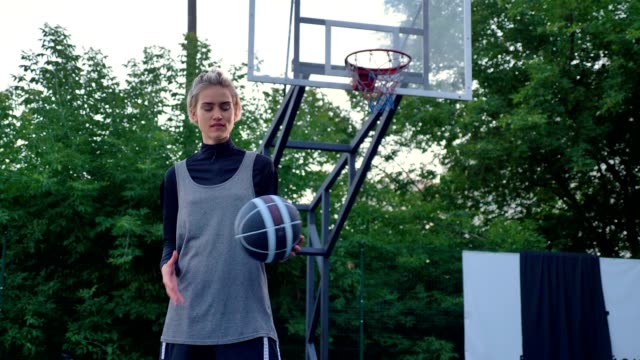 Somebody-throwing-basketball-to-female-player,-woman-catching-ball-and-looking-at-camera,-standing-in-park,-hoop-in-background
