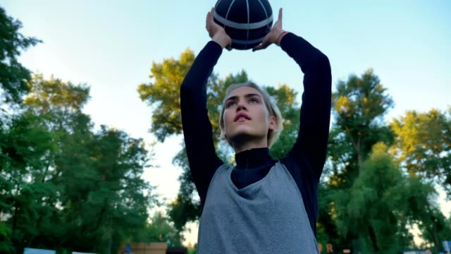 Beautiful-female-basketball-player-throwing-ball,-playing-in-park-with-blue-sky-above,-low-angle