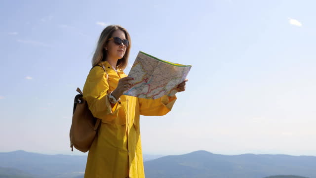 Young-woman-hiking-in-raincoat-on-a-mountain-trail,-stops-and-checks-map-for-directions.-Hiker-reaches-mountain-top,-checks-directions-on-map